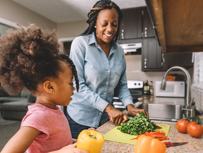 A Black woman chops vegetables while her daughter watches.