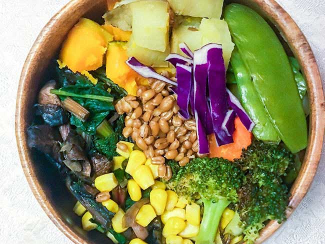 Bowl containing a variety of fresh cooked vegetables including pumpkin, kale, spelt, snow peas, broccoli, corn, red cabbage, and sautéed mushrooms