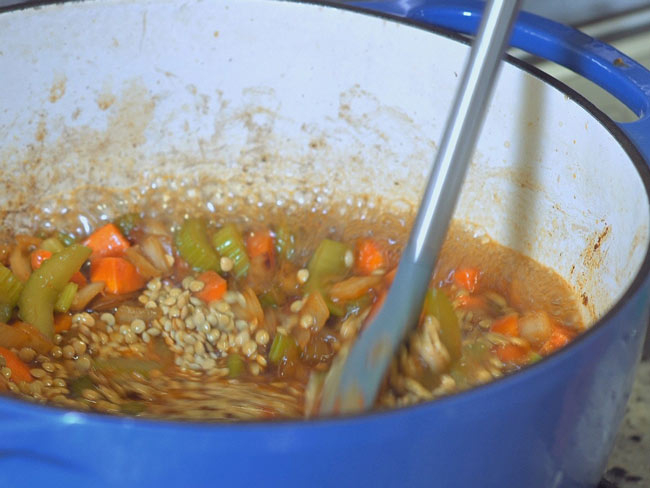 Lentils being mixed with chopped carrots and celery stalks in blue pot.