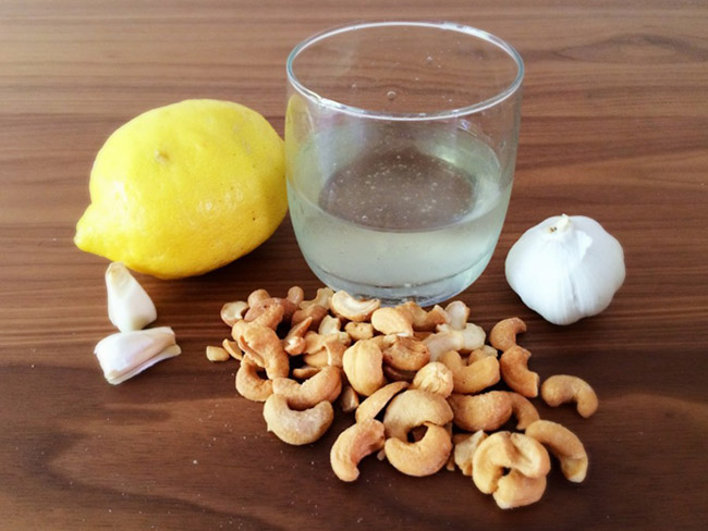 Glass of fresh lemon juice placed next to a garlic bulb, cashews, and a whole lemon